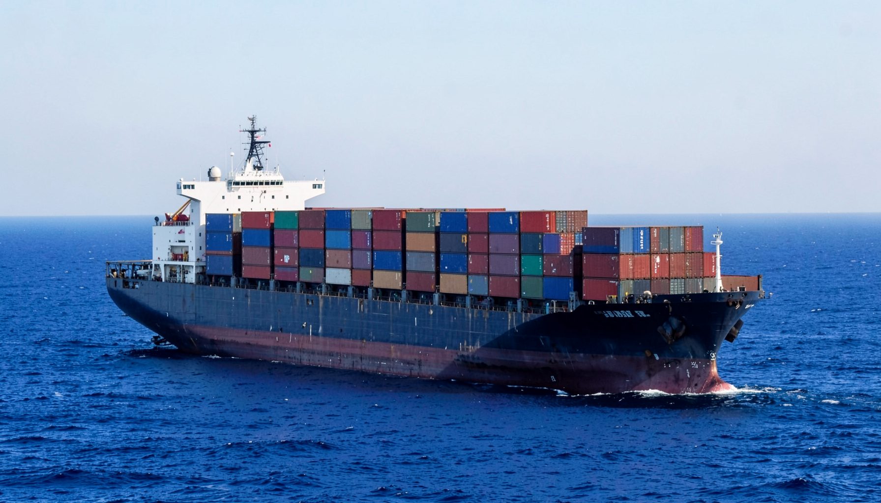 Large cargo ship at sea with stacked shipping containers, deep blue ocean and pale sky, industrial maritime atmosphere