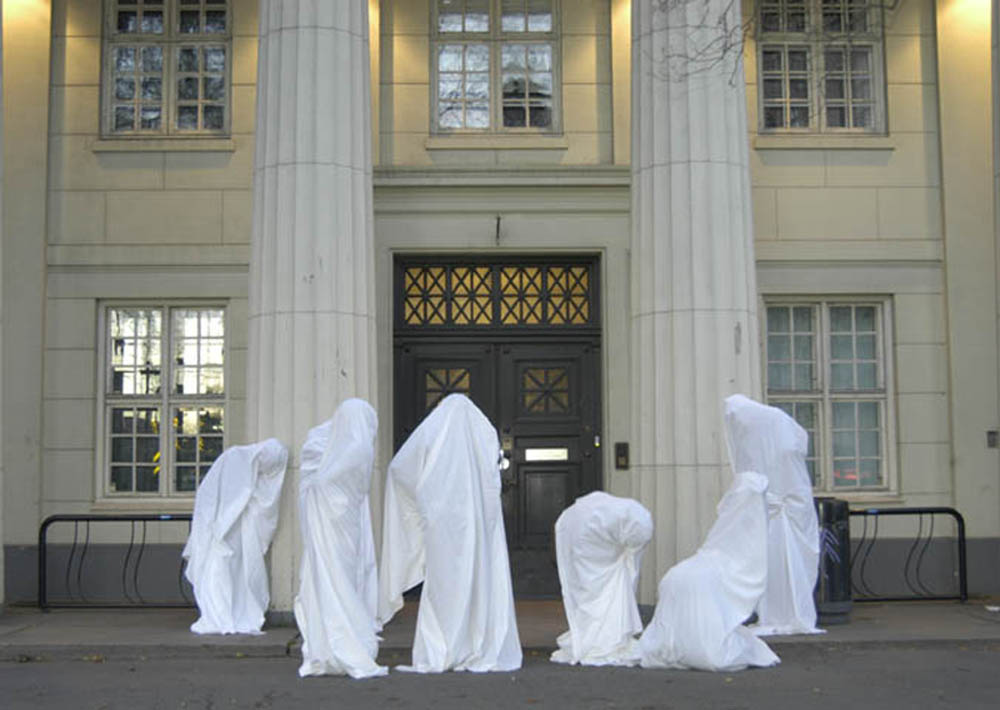 Group of people fully covered in white sheets like ghost figures, posed in front of a classical building facade, muted grey mood
