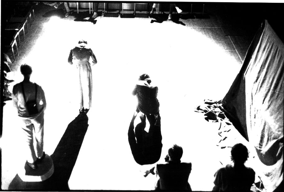 Black and white bright stage with silhouetted figures and a standing model, dramatic high-contrast mood