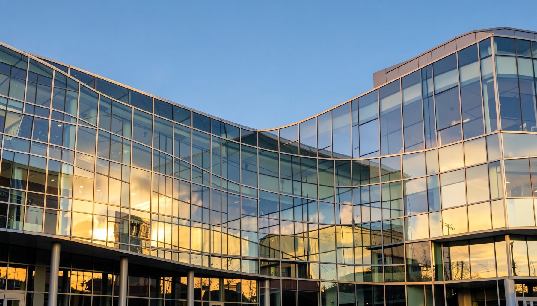 A sweeping view of a modern glass building facade with curved lines, warm golden sunlight reflecting off the windows, set against a clear blue sky